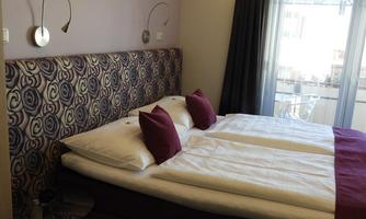 A cozy bedroom with a double bed and a colorful headboard. The room has large windows that lead to a balcony.