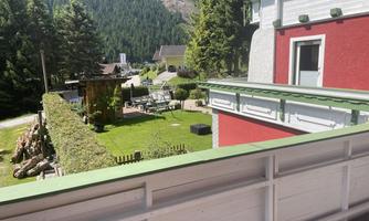 A well-maintained garden with furniture and a green hedge can be seen. In the background, trees and mountains rise.