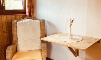 A cozy armchair stands next to a small table against the wall. A picture of a waterfall hangs on the wall, and a window lets in natural light.