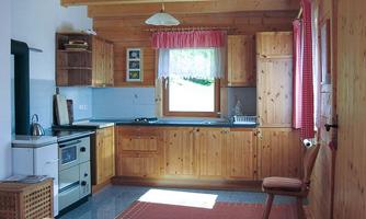 A cozy wooden kitchen with a large window and bright curtains.  
It is well equipped with cooking appliances and a wooden chair.