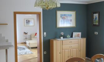 A bright room with a wooden wardrobe, a table, and a colorful lampshade. Through the door, another room with a sofa and a rug is visible.