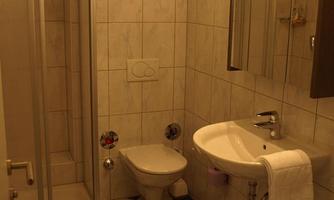 A small bathroom with a shower, sink, and toilet. The walls are tiled, and the floor has a classic pattern.
