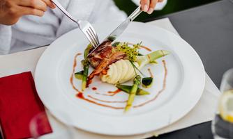 An elegant dish on a white plate, showing meat, mashed potatoes, and vegetables. A person is cutting the food with a knife and fork.