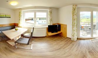 A modern living room with a dining table, black chairs, and a television. Large windows offer a view of the scenery.