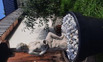 An elegant statue of an implied person sits in a pot with white stones. In the background, there is a wooden wall and some green vegetation visible.