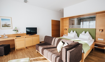 A modern hotel room with a double bed, a television, and a cozy seating area. The furniture is made of light wood, and the room feels welcoming and bright.