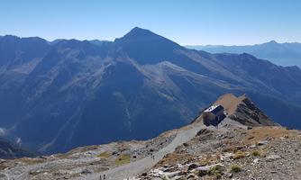 An impressive mountain landscape with high peaks and clear sky. A hiking trail leads to a small cabin near the mountainside.