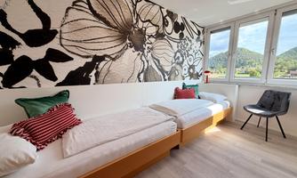 A modern room with two single beds and colorful pillows. An artistic floral pattern can be seen on the wall, and large windows offer a view of nature.