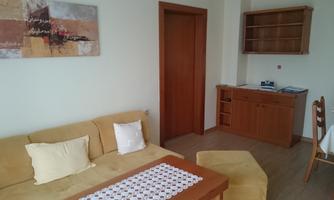 A bright and modern living room with a beige sofa and a small table. In the background, there is a wooden cabinet and a wall painting.