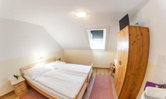 A simple, bright bedroom with two single beds. The room has a wooden wardrobe and a window that provides daylight.
