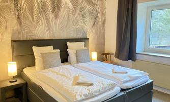 A bright room with two single beds, decorated with a wallpaper of palm leaves. The room has large windows and is equipped with modern lamps.