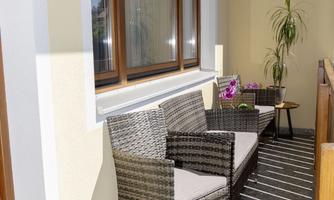 A cozy balcony with two wicker chairs and a small table. In the background, there are plants and plenty of daylight.