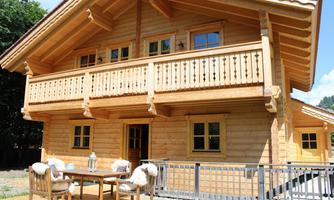A cozy wooden house with a balcony and garden furniture. The facade is made of light wood and there are many windows.