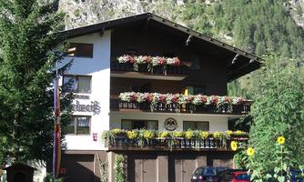 A charming multi-story building adorned with colorful flowers on the balconies. Surrounded by green trees and mountains, it is an inviting place.