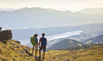Ein Paar steht an einem Berggipfel und schaut auf einen wunderschönen See und die umliegende Landschaft. Die Sonne scheint und die Berge im Hintergrund sind deutlich zu sehen.