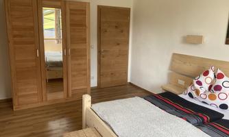 A cozy bedroom with a bed, a large mirror, and a wooden wardrobe. The walls are bright and the floor is made of wood.