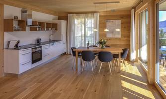 A modern kitchen with wooden surfaces and a large dining table.  
Large windows provide plenty of daylight and a view outside.