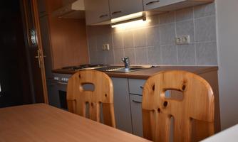 A modern kitchen with wooden chairs and light-colored cabinets. The equipment includes a cooktop, a sink, and functional lighting.