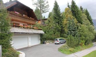 A charming house surrounded by trees and greenery. A car is parked in the driveway and the atmosphere is calm.