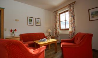 A cozy living room with red sofas and a wooden table. The walls are decorated with pictures, and a window lets in natural light.