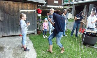 A cheerful gathering outdoors with several people interacting with each other. A woman is dancing while children are watching and standing on the grass.