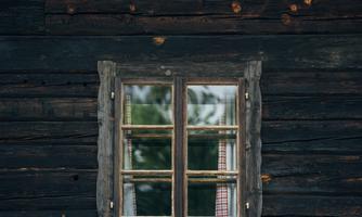 A window with a wooden frame and blooming plants on the sill. The building has a dark facade made of wooden planks.