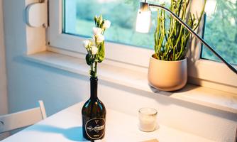 A bright table next to a window, decorated with a bottle of wine, a candle, and a book. In the background, there is a plant in a pot.