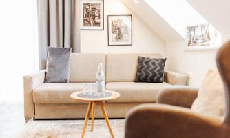 A cozy living room with a beige sofa and a round table. Pictures are hanging on the wall, and the window lets in a lot of light.