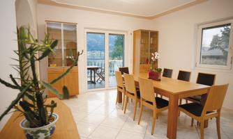 A bright dining area with a large wooden table and chairs. Through the windows, there is a beautiful view of nature.