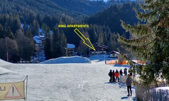 A snow-covered landscape with a ski resort and several people spending time there. In the background, the RMG Apartments are visible among the trees.