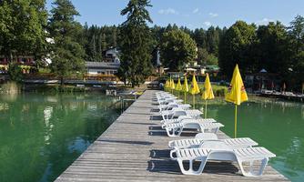 A wooden walkway leads to a tranquil lake with green water. Along the walkway, there are loungers and yellow sun umbrellas.