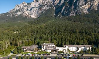 A hotel nestled in a beautiful mountain landscape, surrounded by forests. In the foreground are parking spaces and a clear view of the mountains.
