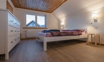 A bright bedroom with a double bed, a dresser, and two wall lamps. Large windows offer a view outside and provide daylight.