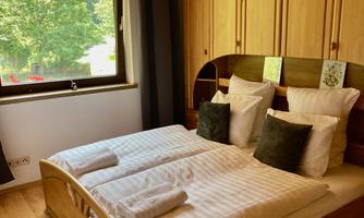 A bright bedroom with a double bed and beige cushions. Large windows offer a view of the green surroundings.