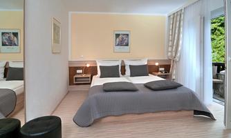 A modern bedroom with a cozy double bed and soft pillows. Large windows provide plenty of daylight and a view of the greenery.