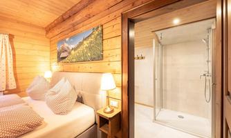 A cozy room with wooden paneling and a large bed. In the background, there is a modern bathroom with a shower.