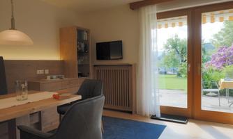 A bright living space with a table, chairs, and a television. Large windows offer a view of the garden and the nature outside.