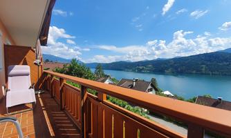 A beautiful balcony with a view of a clear lake and surrounding mountains. The sky is blue with some clouds.
