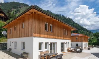 Two modern wooden houses in a picturesque mountain landscape. The buildings feature terraces with seating and are surrounded by green trees.