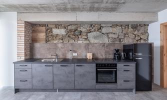 A modern kitchen with gray furniture and a concrete wall accent. The equipment includes a stove, refrigerator, and coffee machine.