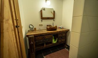 A modern bathroom with a rustic wooden washbasin and a mirror. The walls are light, and the floor is dark tiled.