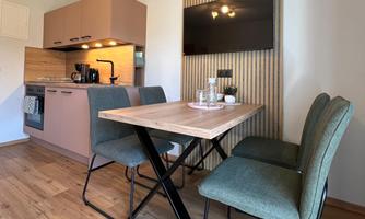A modern kitchen unit with a dining table and four chairs. The room is bright and friendly with wooden flooring and a wall with a television.