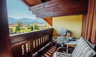 A cozy balcony with two chairs and a table, set with breakfast. In the background, green meadows and mountains can be seen.