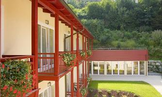 A modern building with red balconies and flower boxes. The background shows a green, wooded landscape.