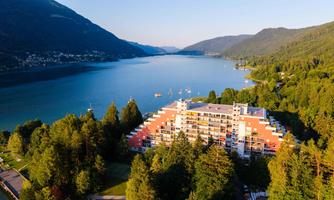 A modern hotel by the shore of a clear lake, surrounded by lush forests and mountains. The tranquil landscape invites relaxation.