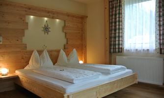 A cozy bedroom with a wooden wardrobe and two beds. The room has large windows with curtains and a warm, inviting atmosphere.