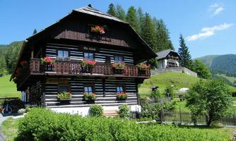 A traditional wooden house with flowers on the balcony in an idyllic landscape. In the background, green hills and trees can be seen.