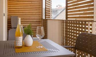 An inviting balcony with a table, a bottle of wine, and a glass. In the background, modern wooden elements and a beautiful view can be seen.