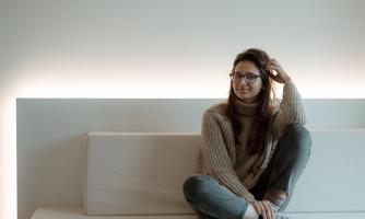 A young woman is sitting on a light couch in a minimalist room. Soft light is gently shining from the wall behind her.