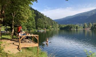 A beautiful lake surrounded by trees and mountains. One person is standing in the water while others are sitting on the shore and relaxing.